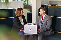 In einer Sitzecke in einem Unternehmen spricht eine Vertreterin des DRK mit einem Mann im Anzug über die verschiedenen Angebote des DRK