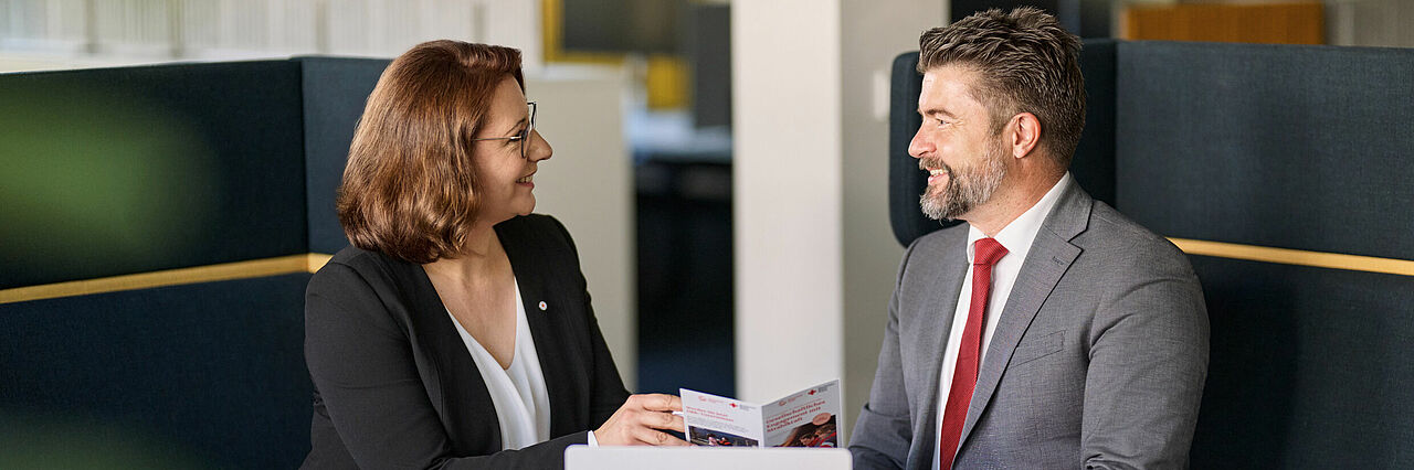 In einer Sitzecke in einem Unternehmen spricht eine Vertreterin des DRK mit einem Mann im Anzug über die verschiedenen Angebote des DRK